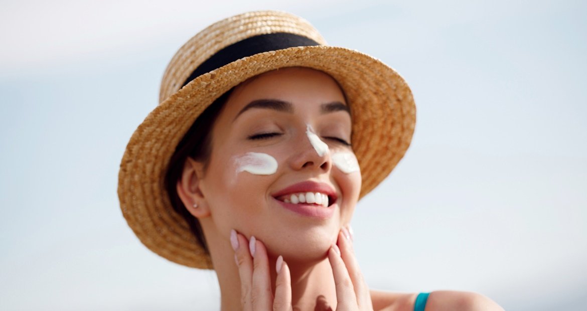 Mulher branca ao ar livre sorri enquanto aplica o protetor solar para proteger a pele e mantê-la saudável.